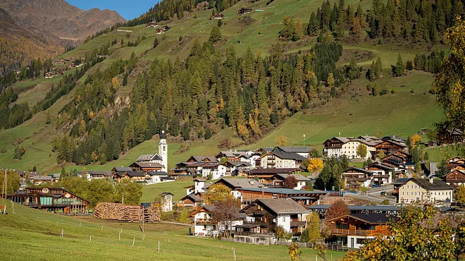 Ein malerisches Bergdorf in einer alpinen Landschaft. Im Mittelgrund liegen traditionelle Holzhäuser mit spitz zulaufenden Dächern, daneben ein Kirchturm mit einer Uhr. Die Gebäude sind umgeben von grünen Wiesen und Waldflächen, während im Hintergrund bewaldete Berghänge zu sehen sind. Die herbstlichen Farben der Bäume deuten auf die Jahreszeit hin.
