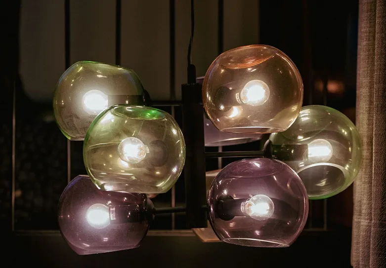 Eine moderne Deckenlampe mit mehreren kugelförmigen Glühbirnen, die in metallischen Fassungen angeordnet sind. Die Glühbirnen sind in verschiedenen transparenten Farben, wie lila, grün und braun, gehalten und strahlen warmes Licht aus.