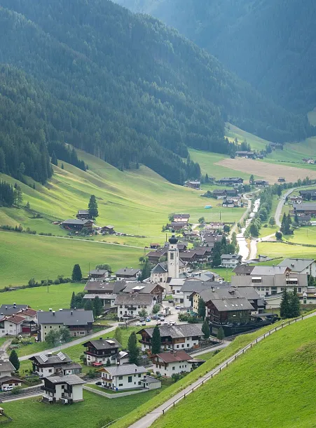 Ein malerisches Tal mit einem kleinen Dorf inmitten grüner Hügel und bewaldeter Berge. Die Szene zeigt eine verstreute Ansiedlung von Häusern, asphaltierte Straßen und eine zentrale Kirche. Die Umgebung ist üppig grün mit Wiesen und Bäumen, die sich in sanften Hügeln und dichten Wäldern erstrecken. Der Himmel ist teilweise von Wolken bedeckt, wodurch ein harmonisches und ruhiges Landschaftsbild entsteht.