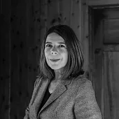 Eine schwarz-weiße Fotografie zeigt eine Frau mit schulterlangem Haar, die in die Kamera lächelt. Sie trägt ein Jackett über einem Oberteil. Der Hintergrund scheint eine Holzwand zu sein.