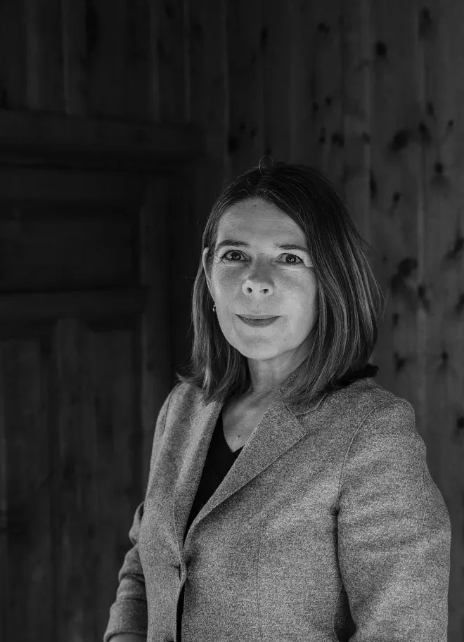 Portrait einer Person in Schwarz-Weiß, die einen Blazer trägt. Die Person hat mittellanges Haar und schaut direkt in die Kamera. Der Hintergrund ist unscharf und zeigt vermutlich Holzwände oder -paneele.