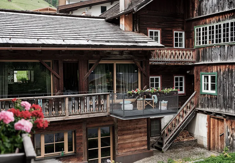 Ein traditionelles alpines Holzhaus mit einem steilen Schrägdach und mehreren Balkonen. Der Vordergrund zeigt blühende Blumen in einem Blumenkasten. Im Hintergrund sind weitere hölzerne Gebäudeteile und grüne Hügel zu sehen.