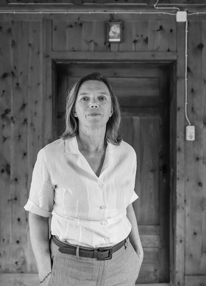 Eine Frau in einem weißen Hemd steht vor einer Holzwand. Sie hat schulterlanges Haar und sieht direkt in die Kamera. Die Umgebung wirkt rustikal mit Holzverkleidungen und einer Tür im Hintergrund.