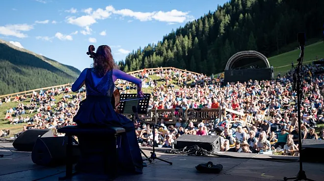 Eine Musikerin spielt Cello auf einer Bühne im Freien, während ein großes Publikum auf einer Wiese im Hintergrund sitzt. Der Himmel ist klar, und die Landschaft ist von bewaldeten Hügeln umgeben.