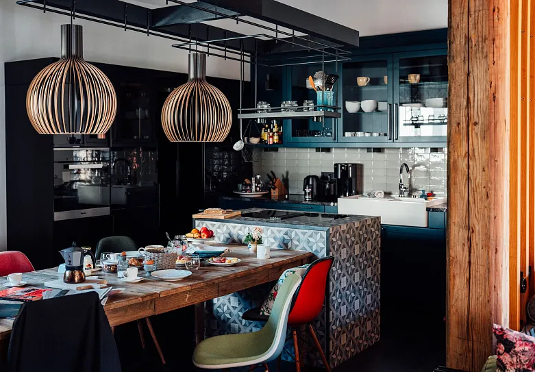 Ein modernes Küchen- und Esszimmer mit einem Holztisch, an dem mehrere bunte Stühle stehen. Die Küche ist mit dunklen Schränken ausgestattet, und auf dem Tisch steht Geschirr und verschiedene Gegenstände. Zwei große hängende Lampen sind auffällig über dem Tisch angebracht. Die Wand hat eine weiße Kachelrückwand hinter der Spüle und verschiedene Küchenutensilien sind sichtbar.