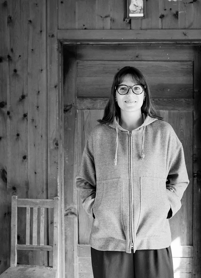Eine Person mit Brille und einem Kapuzenpullover steht in einem Raum mit Holzwänden und einem Holzstuhl. Die Person lächelt, die Hände in den Taschen des Pullovers.