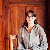 Eine Person sitzt auf einem Stuhl vor einer Holzwand. Die Person trägt eine Brille und einen grauen Kapuzenpullover. Der Hintergrund besteht aus einer gestrichenen Holzwand.