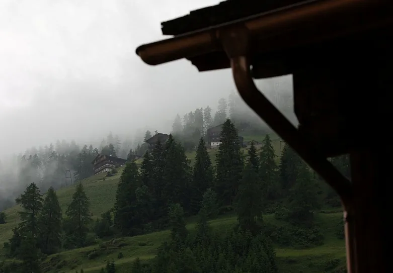 Eine neblige Berglandschaft mit mehreren Holzhäusern am Hang, umgeben von dichten Nadelwäldern.