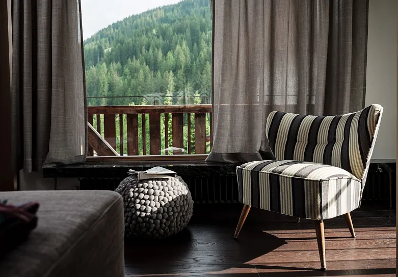 Ein gemütliches Wohnzimmer mit einem gestreiften Sessel neben einem großen Fenster, das den Blick auf eine bewaldete Berglandschaft freigibt. Vor dem Fenster steht ein kleiner, gestrickter Hocker.