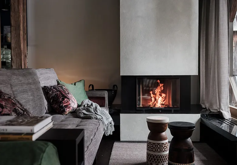 Gemütliches Wohnzimmer mit einem modernen Kamin, in dem ein Feuer brennt. Es gibt ein graues Sofa mit mehreren Kissen in verschiedenen Farben, darunter Grün und Rosa. Vor dem Sofa stehen zwei dekorative Hocker. Der Raum hat eine warme und einladende Atmosphäre mit einem braunen Holzfußboden und einem Teppich.