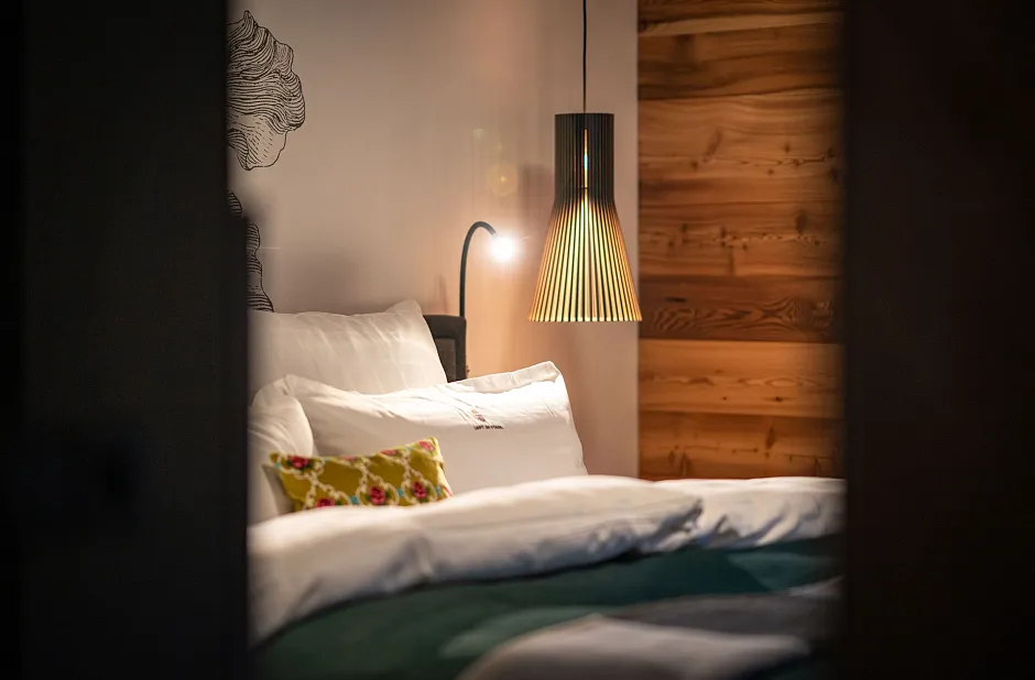 Ein gemütliches Schlafzimmer mit weißer Bettwäsche und grüner Decke. An einer Wand hängt eine moderne Lampe und im Hintergrund ist eine Wand mit Holzverkleidung zu sehen. Ein dekoratives Kissen mit Blumenmuster liegt auf dem Bett.