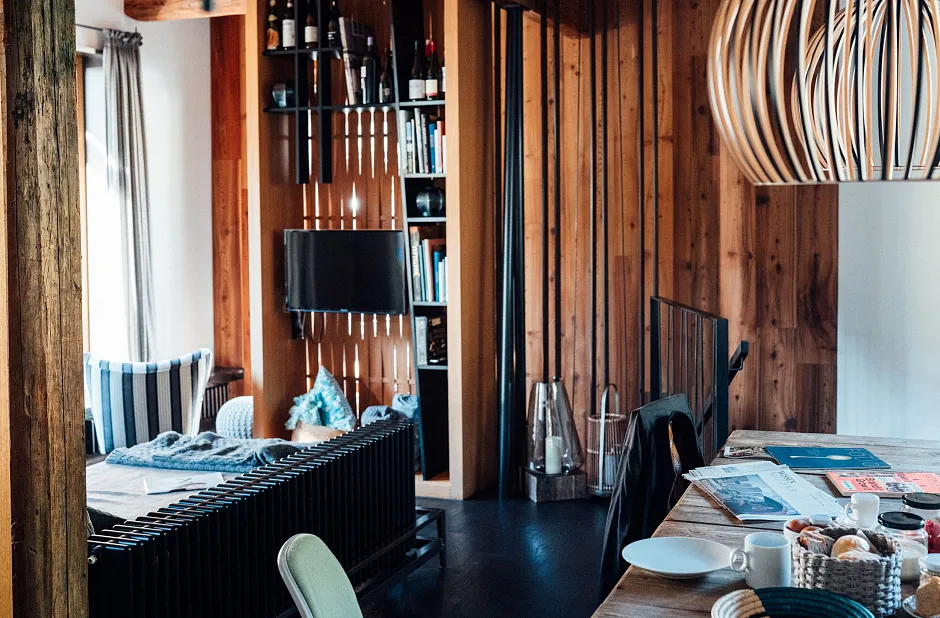 Gemütliches Wohnzimmer mit Holzwänden, einem Regal mit Weinflaschen und Büchern, einem kleinen Fernseher, einem Sofa mit gestreiften Kissen und einem Tisch mit Geschirr und einer Zeitung.