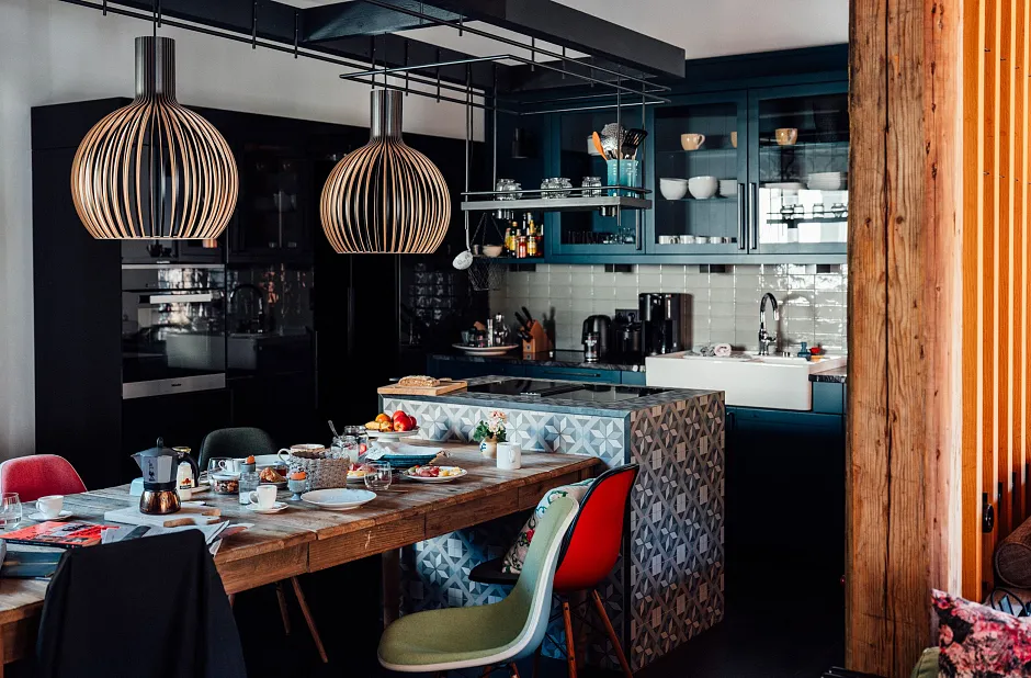 Ein modernes Küchen- und Esszimmer mit einem Holztisch, an dem mehrere bunte Stühle stehen. Die Küche ist mit dunklen Schränken ausgestattet, und auf dem Tisch steht Geschirr und verschiedene Gegenstände. Zwei große hängende Lampen sind auffällig über dem Tisch angebracht. Die Wand hat eine weiße Kachelrückwand hinter der Spüle und verschiedene Küchenutensilien sind sichtbar.