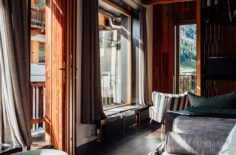 Ein gemütlicher Wohnraum mit großen Fenstern, die viel Tageslicht hereinlassen. Im Raum befinden sich ein graues Sofa und Sessel, und die Wände sind aus Holz. Durch die Fenster sieht man einen Balkon und eine Berglandschaft im Hintergrund.