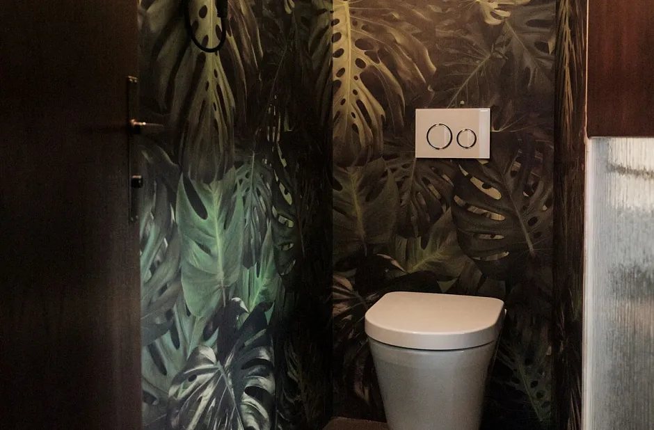 Ein Badezimmer mit Tapeten im Dschungelmotiv. Die Wände sind mit Blättern in Grüntönen bedeckt. Es gibt eine moderne Toilette mit einem zweistufigen Spülknopf und einen kleinen Wandleuchter über der Tür.
