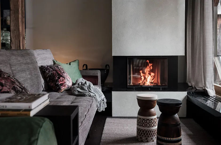 Gemütliches Wohnzimmer mit einem modernen Kamin, in dem ein Feuer brennt. Es gibt ein graues Sofa mit mehreren Kissen in verschiedenen Farben, darunter Grün und Rosa. Vor dem Sofa stehen zwei dekorative Hocker. Der Raum hat eine warme und einladende Atmosphäre mit einem braunen Holzfußboden und einem Teppich.