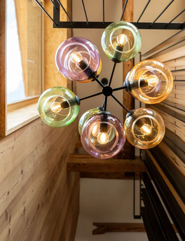 Eine Deckenleuchte mit mehreren bunten Glaskugeln, die von oben betrachtet wird, hängt in einer Innenumgebung mit Holzwänden und Holzdecke. Die Glaskugeln haben verschiedene Farben wie Grün, Lila und Gelb und sind um Glühbirnen angeordnet. An den Seiten sind Holzwände und schwarze Metallgitter zu sehen, die Teil eines Treppengeländers sein könnten.