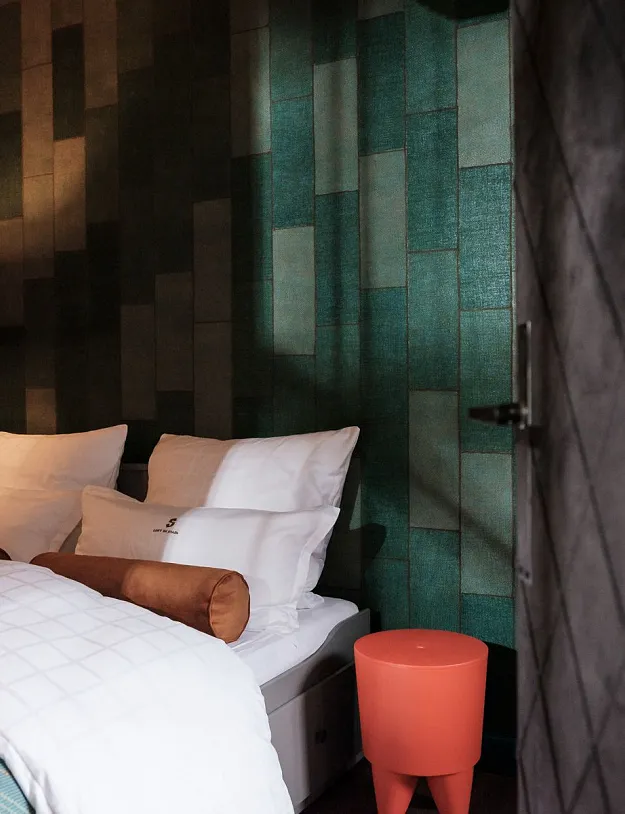 Ein stilvoll eingerichtetes Schlafzimmer mit einem Bett, das mit weißen Kissen und einem braunen zylinderförmigen Kissen dekoriert ist. Neben dem Bett steht ein roter, kleiner Beistelltisch. Die Wände haben eine moderne, gemusterte Verkleidung in Erdtönen, und eine Lampe mit einem rechteckigen Schirm spendet warmes Licht.