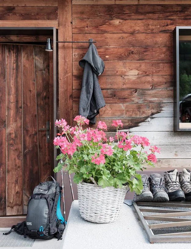Eine rustikale Holzhütte mit einem Fenster und blühenden rosa Geranien in einem Korb davor. Neben der Tür hängt eine Jacke, und auf dem Boden stehen ein Rucksack und Wanderstiefel. Im Hintergrund sind Berge zu sehen.
