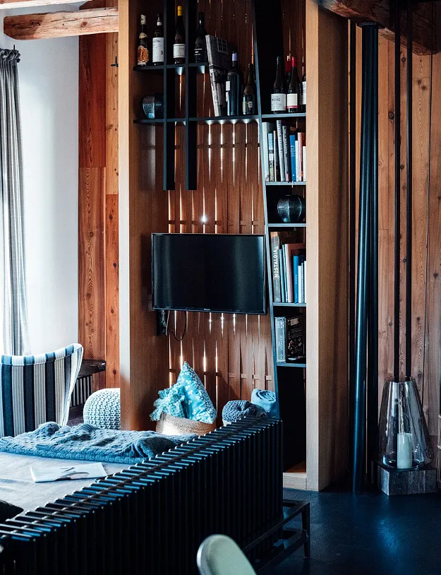 Ein gemütlicher Raum mit Holzwänden und moderner Einrichtung. Ein Fernseher ist an einer Wand befestigt, und es gibt Regale mit Büchern und Weinflaschen. Ein Fenster mit Vorhängen lässt natürliches Licht herein. Vor dem Fernseher steht ein Sofa mit dekorativen Kissen.
