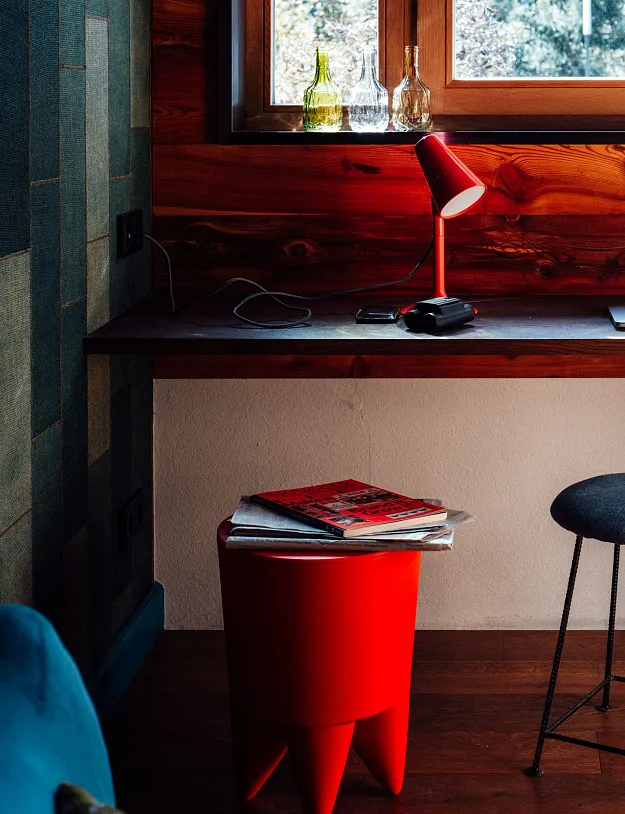 Ein Raum mit einem Schreibtisch vor einem Fenster, das Tageslicht hereinlässt. Auf dem Tisch steht eine rote Schreibtischlampe, daneben liegt ein Laptop. Auf dem Boden steht ein roter Hocker mit einer Zeitschrift darauf. Rechts von der Lampe befindet sich ein geflochtener Hocker. Die Wände sind teils aus Holz und teils in einem dunklen Grünton gehalten.