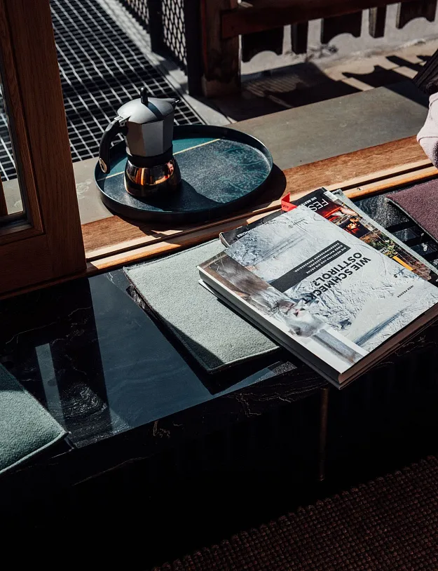 Ein gemütliches Wohnambiente mit einem Fensterbrett, auf dem ein Magazin, ein Buch, eine Kanne und Stoffe liegen. Draußen sind Holzgeländer und Sonnenlicht zu sehen.
