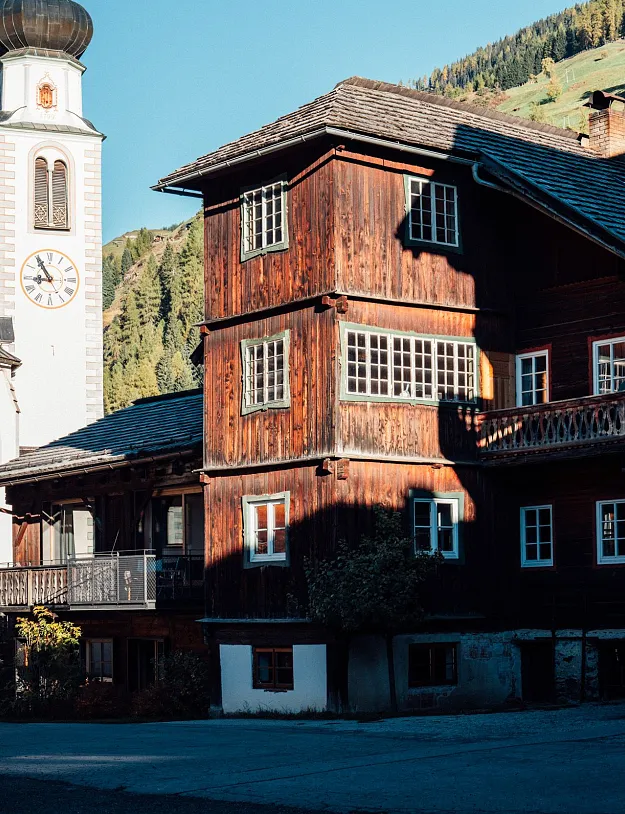 Ein malerisches Bergdorf mit einer Kirche und traditionellen Holzhäusern im Vordergrund. Die Sonne beleuchtet Teile der Gebäude, während die umliegenden Berge einen idyllischen Hintergrund bilden.