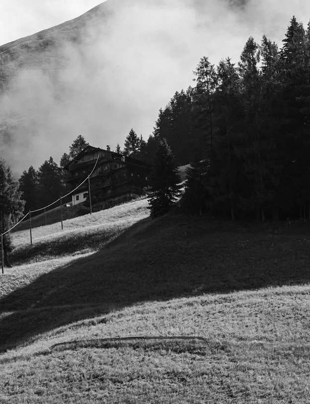 Eine schwarz-weiße Landschaftsaufnahme zeigt eine Berghütte, umgeben von Bäumen. Ein steiler Hang führt zur Hütte hinauf, während im Hintergrund ein bewaldeter Berg zu sehen ist und Wolken den Himmel bedecken.