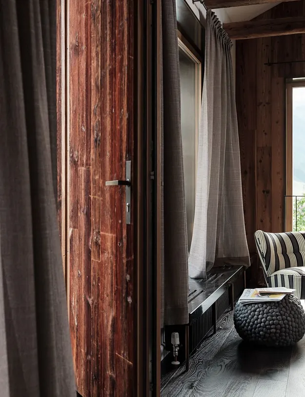 Ein gemütlicher Innenraum mit Holzwänden und -böden. Im Bild gibt es einen Stuhl mit gestreiftem Stoff und ein kleines geflochtenes Beistelltischchen. Große Fenster mit hellen Vorhängen lassen viel Tageslicht herein und bieten einen Ausblick auf grüne Berge.