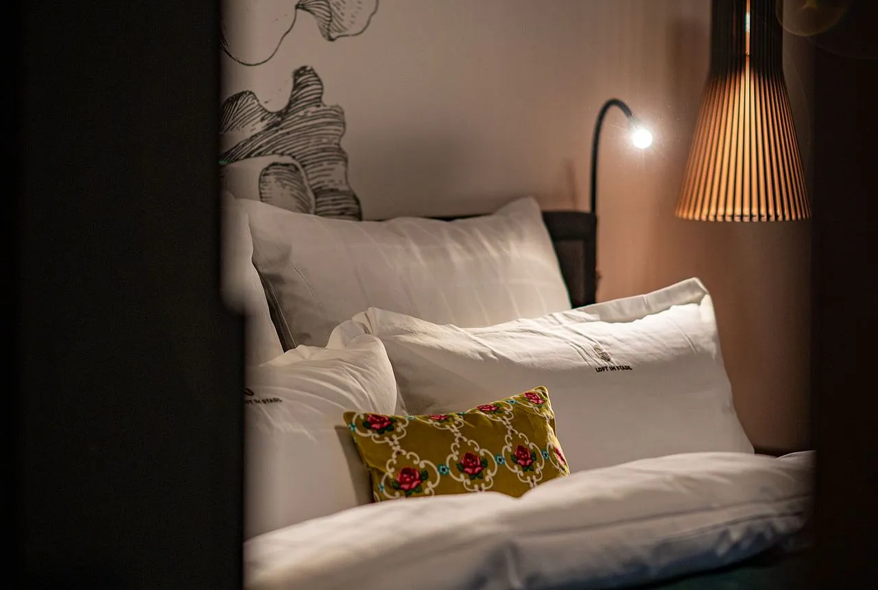 Ein gemütliches Schlafzimmer mit weißen Kissen und Bettwäsche, dekoriert mit einem kleinen, gemusterten Kissen in der Mitte. Im Hintergrund ist eine stilvolle Lampe zu sehen, die warmes Licht abgibt, sowie eine Wand mit einem floralen Muster.