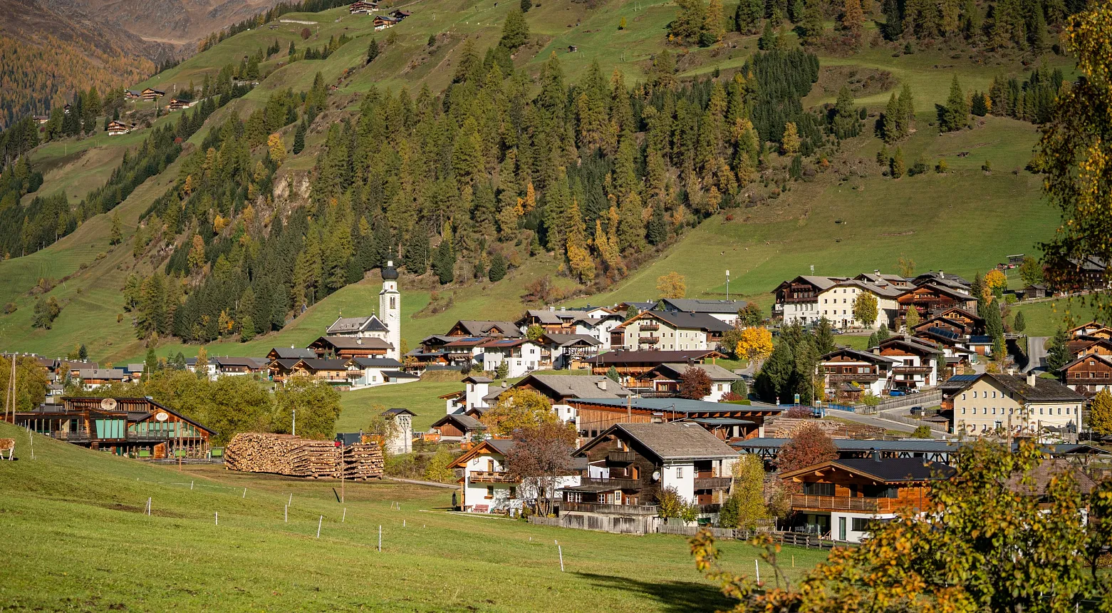Ein malerisches Bergdorf in einer alpinen Landschaft. Im Mittelgrund liegen traditionelle Holzhäuser mit spitz zulaufenden Dächern, daneben ein Kirchturm mit einer Uhr. Die Gebäude sind umgeben von grünen Wiesen und Waldflächen, während im Hintergrund bewaldete Berghänge zu sehen sind. Die herbstlichen Farben der Bäume deuten auf die Jahreszeit hin.