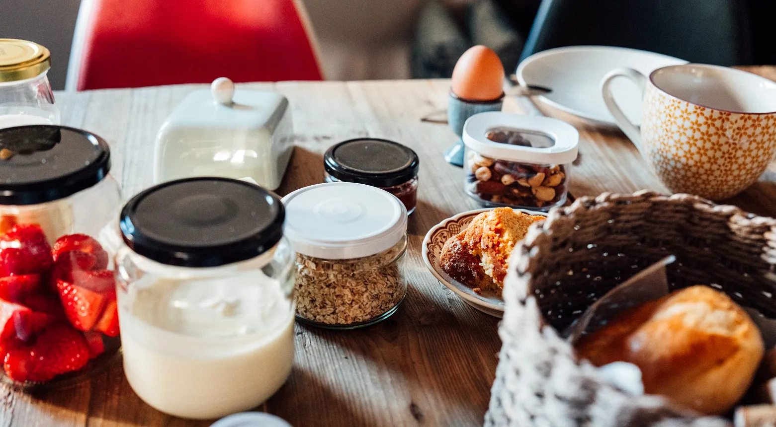 Ein Frühstückstisch mit verschiedenen Lebensmitteln. Auf dem Tisch stehen Gläser mit Joghurt, Erdbeeren, Haferflocken und Nüssen. Es gibt auch ein weichgekochtes Ei in einem Eierbecher, einen Korb mit Brötchen und eine Tasse. Der Tisch ist aus Holz.
