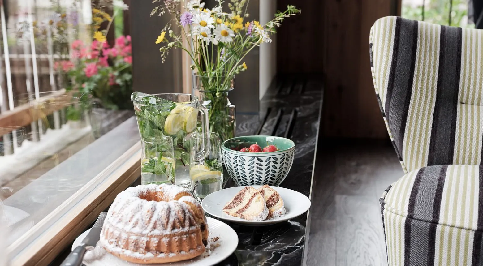 Auf einer Fensterbank steht ein gedeckter Tisch mit einem Kuchen und einer Glasschale mit Gurkenscheiben im Wasser. Daneben befindet sich eine Schüssel mit Müsli oder Joghurt und ein Teller mit Brotscheiben. Ein Blumenstrauß in einer Vase schmückt den Tisch. Im Hintergrund sind grüne Pflanzen und bunte Blumen zu sehen.