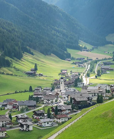 Ein malerisches Tal mit einem kleinen Dorf inmitten grüner Hügel und bewaldeter Berge. Die Szene zeigt eine verstreute Ansiedlung von Häusern, asphaltierte Straßen und eine zentrale Kirche. Die Umgebung ist üppig grün mit Wiesen und Bäumen, die sich in sanften Hügeln und dichten Wäldern erstrecken. Der Himmel ist teilweise von Wolken bedeckt, wodurch ein harmonisches und ruhiges Landschaftsbild entsteht.